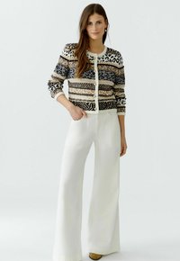 Strickjacke mit gemischten Mustern in Schwarz, Beige und Weiß, mit Rundhalsausschnitt und Knopfverschluss, kombiniert mit weiten weißen Hosen.