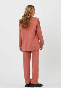 Conjunto de dos piezas rosa hecho de tejido tejido. La chaqueta tiene un corte cuadrado y los pantalones son rectos. Zapatos negro completan el look.