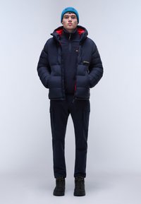 Marineblauer Pufferjacke mit roter Innenausstattung, marineblauer Fleece darunter und Utility-Hosen. Schwarze wasserdichte Stiefel; türkisfarbene Mütze. Frontale Pose vor weißem Hintergrund.