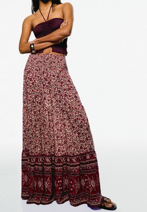 Femme portant un haut licou bordeaux et une longue jupe à motifs floraux avec une large bordure rouge, debout les bras croisés.