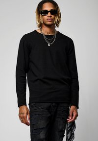 Maglietta nera a maniche lunghe con scollatura rotonda, abbinata a jeans neri strappati. Gli accessori includono più catene d'argento e occhiali da sole.