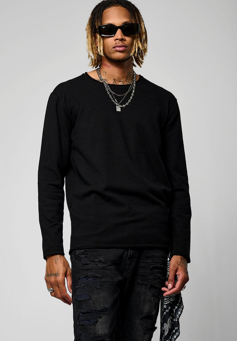 Maglietta nera a maniche lunghe con scollatura rotonda, abbinata a jeans neri strappati. Gli accessori includono più catene d'argento e occhiali da sole.