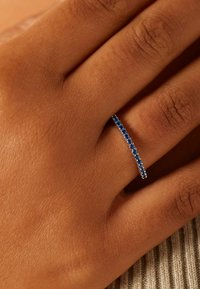Zilveren ring met een rij kleine blauwe stenen, gedragen aan een hand met een getextureerde, gebruinde huid. Eenvoudig, slank ontwerp.