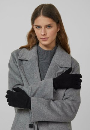 Jonge vrouw draagt een grijze jas, grijze trui en zwarte handschoenen, met haar armen over elkaar, tegen een effen grijze achtergrond.