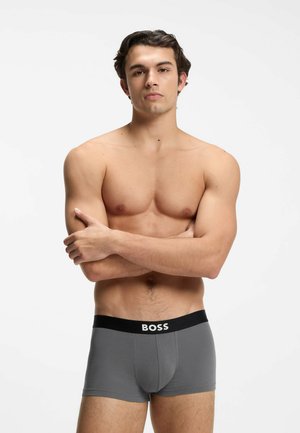 Skjorteløs mand med krydsede arme iført grå boksertrusser med sort linning mærket "BOSS" mod en ensfarvet hvid baggrund.