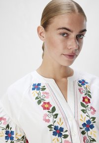 Blusa bianca ricamata con motivi floreali in blu, rosa e giallo, con chiusura a bottoni e colletto alla cinese.
