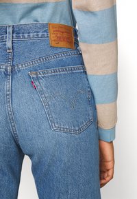 Jeans de mezclilla azul de cintura alta, con un parche de cuero marrón y una etiqueta roja icónica en el bolsillo trasero, que muestran un diseño clásico.