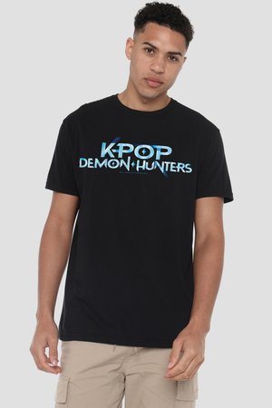 Junger Mann mit kurzen lockigen Haaren, der ein schwarzes T-Shirt mit blauem Text "K-POP DEMON HUNTERS" und beige Cargoshorts vor grauem Hintergrund trägt.