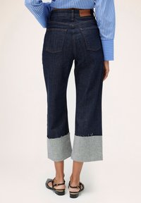 Pantaloni largi din denim închis, cu un tiv gri scurtat, margini destramate și o etichetă de piele branduită pe talie.