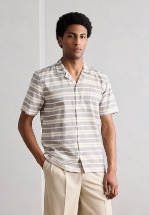 Νεαρός άνδρας με κοντά σγουρά μαλλιά, φορώντας κοντομάνικο γκρι και λευκό ριγέ πουκάμισο με κουμπιά και μπεζ παντελόνι, στέκεται με το ένα χέρι στην τσέπη.