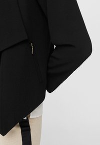 Veste noire au design superposé, avec une poche latérale zippée et un tissu texturé. L'ourlet est asymétrique et incliné.