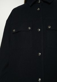 Veste en laine bleu marine foncé avec boutons-pression argentés, comprenant deux poches poitrine à rabats boutonnés et un col pointu.