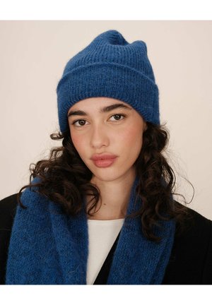 Bonnet tricoté bleu avec une texture côtelée et un revers. Porté avec une écharpe bleue assortie, toutes deux en fil doux et pelucheux.