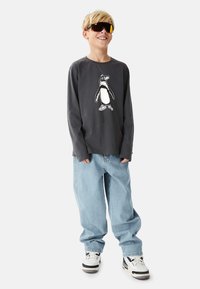 Graues Langarmshirt mit Pinguin-Motiv, hellblaue weite Jeans und weiße Turnschuhe. Accessoires umfassen große schwarz-gelbe Sonnenbrillen.