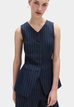 Kobieta w granatowej kamizelce w prążki bez rękawów z pięcioma guzikami z przodu i dopasowanych spodniach, stoi na tle gładkiego tła.