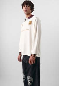 Crèmekleurig polo shirt met lange mouwen en een bordeaux kleurige kraag, voorzien van geborduurde logo's en tekst, gecombineerd met donkere spijkerbroeken met witte boomontwerpen.