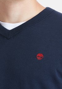 Tmavě modrý svetr s výstřihem do V vyrobený z měkké pleteniny, s malým červeným logem na levé straně hrudi. Klasický design s žebrovanými lemy.