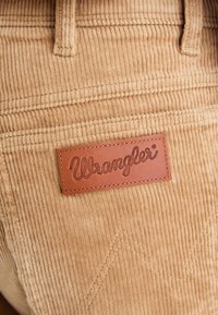 Hnědá kordurová látka s vertikálním pruhem. Obsahuje hnědou koženou nášivku Wrangler. Přítomno jemné šití do diamantového vzoru.