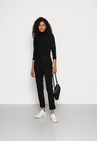 Pull à col roulé noir, pantalon noir ajusté, baskets blanches et un petit sac noir. Le mannequin se tient devant un fond blanc uni.