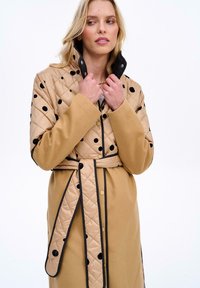 Prošívaný béžový kabát s černými puntíky, odnímatelným límcem bundy a odpovídajícím opaskem, s černým lemováním podél švů.