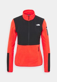Giacca in pile rosso e nero con colletto alto, zip a metà davanti e pannelli neri su spalle e maniche; presenta un logo del marchio e una tasca laterale.