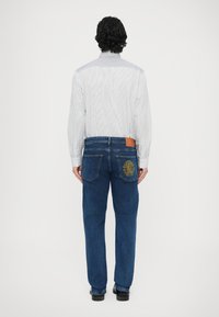 Camisa de botones a rayas con detalles grises, combinada con vaqueros azules que presentan un detalle bordado amarillo en los bolsillos traseros. Zapatos negros visibles.