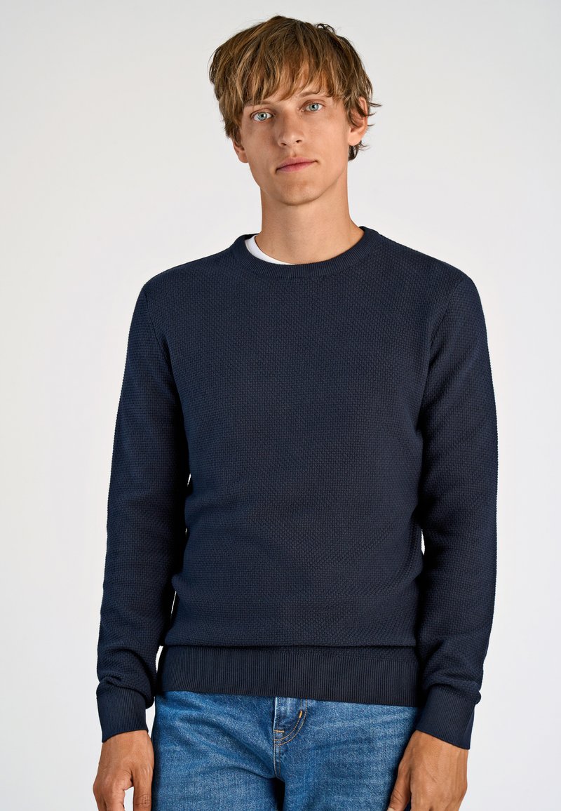 Navyblaue Strickpullover mit strukturierter Oberfläche, Rundhalsausschnitt und gerippten Bündchen sowie Saum, kombiniert mit hellblauen Jeans.