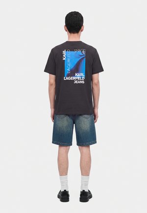 Мъж, носещ черна тениска Karl Lagerfeld Jeans с син графичен дизайн и надпис на гърба, дънкови къси панталони, бели чорапи и черни обувки, стои с гръб към нас.