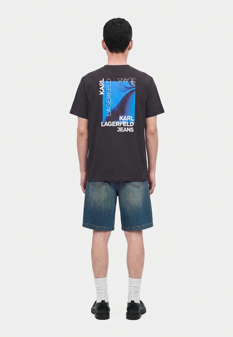 Άνδρας φοράει μαύρο t-shirt Karl Lagerfeld Jeans με μπλε γραφικά και κείμενο στην πλάτη, τζιν σορτς, λευκές κάλτσες και μαύρα παπούτσια, στέκεται με την πλάτη προς τα εμπρός.