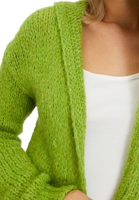 Cardigan verde a maglia con vestibilità ampia, caratterizzato da una finitura testurizzata e un design aperto, indossato sopra una maglietta bianca.