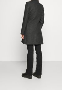 Cappotto di lana grigio con un design attillato, orlo svasato e dettagli di cucitura visibili. Abbinato a pantaloni neri e stivali neri robusti.