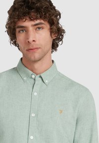Lichtgroene button-up blouse van gestructureerde stof, met een kraag, witte knopen en een klein, oranje geborduurd logo op de borst.
