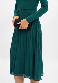 Turkusowa plisowana sukienka z długimi rękawami, z dopasowaną górą i rozkloszowaną spódnicą, trzymana w luźnym uścisku z czarną kopertówką.