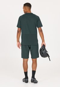 Donkergroen sportoutfit voor mannen bestaande uit een shirt met korte mouwen met textuur en shorts, met een zwarte helm in de linkerhand.