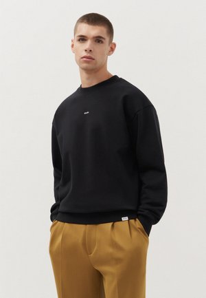 Felpa nera con scollo a girocollo e logo "studio" ricamato sul petto; abbinata a pantaloni beige plissettati.