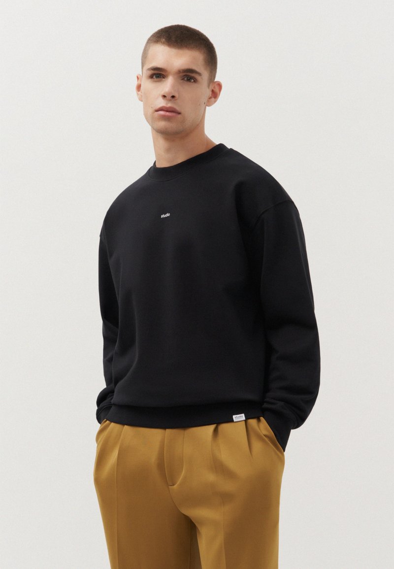 Felpa nera con scollo a girocollo e logo "studio" ricamato sul petto; abbinata a pantaloni beige plissettati.