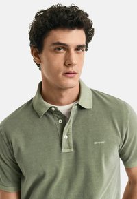 Olivgrünes Polohemd aus Baumwolle mit einem klassischen Kragen, einer zwei-knöpfigen Knopfleiste und einer weichen Textur, mit einem kleinen Logo auf der Brust.