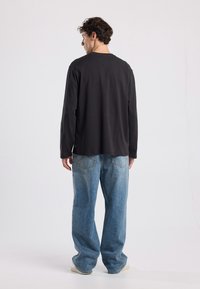 Black long-sleeve shirt with a relaxed fit, paired with light blue wide-leg jeans and beige sneakers. Plain back view, minimal detailing.