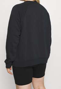 Svart sweatshirt med raglanärmar och ribbad nederkant, tillverkad av mjukt tyg. Bärs med svarta shorts, vilket visar en avslappnad passform.