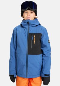 Un adolescent portant une veste d'hiver bleue et noire, un pantalon orange, des gants noirs, un bonnet noir et des lunettes de ski sombres sur la tête, faisant face à l'avant.