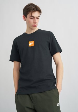 Mladi moški z rjavimi lasmi, oblečen v črno majico s svetlo oranžnim logotipom Nike in zelenimi kratkimi hlačami Nike, z rokami v žepih, gleda navzdol desno.