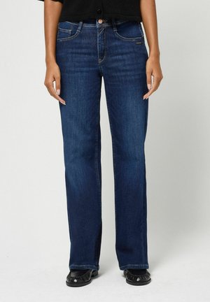 Frau trägt dunkelblaue Straight-Leg-Jeans mit fünf Taschen und schwarzen Schuhen vor einem schlichten hellen Hintergrund.