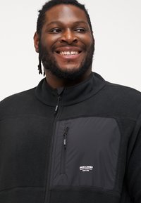 Black fleece jacket with a high collar, zip-up front, and a contrasting black nylon chest pocket featuring a white logo.