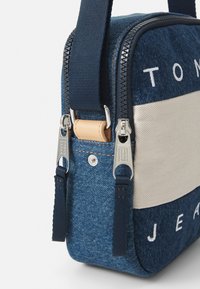 Denim crossover-veske med beige stripe, sølvglidelåser og marineblå detaljer. Den har et teksturert stoff og justerbar stropp.