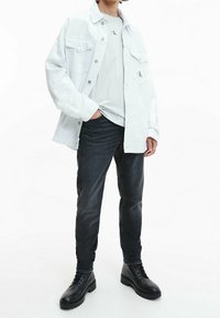 Vit oversized jeansjacka, grå T-shirt med logotyp, mörka jeans och svarta snörstövlar. Slätt tyg, avslappnad passform och modern design.