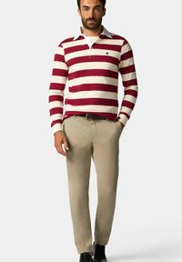 Polo s dlouhým rukávem, červeno-bílými pruhy a límečkem, spárovaný s hnědými kalhotami a černými botami. Na hrudi jednoduché logo.