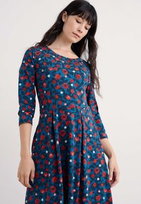 Kukkamekko tummansinisellä pohjalla, jossa on punaisia ja valkoisia kukkia. Pitkät hihat, pyöreä pääntie ja levenevä helma. Pehmeä, teksturoitu kangas.