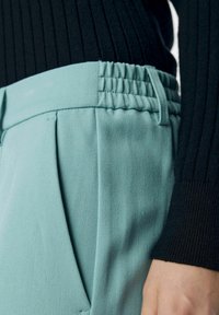 Ljusblå byxor med texturerad midjeband, med veck och sidofickor. En svart ribbad tröja är delvis synlig.