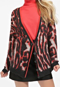 Černo-červený vzorovaný kardigan s chlupatou texturou, otevřeným předním dílem a čtyřmi černými knoflíky. Nošený přes červený rolák.