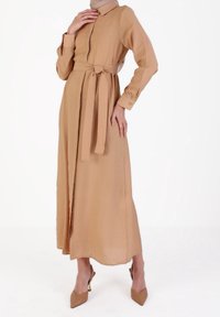 Kvinna dragen i en beige långärmad klänning med bälte, matchande högklackade skor och en beige huvudduk, som står mot en vit bakgrund.